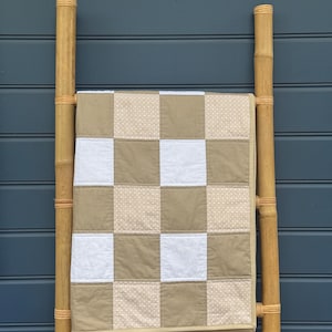 May include: A patchwork quilt in a checkerboard design of white, beige and dotted beige squares. The quilt is displayed on a bamboo ladder against a blue wall. A cosy home decor item.