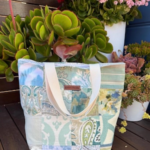 May include: A patchwork tote bag in shades of blue, green, and cream. It features white handles and a small brown leather tag. The bag is displayed on a wooden surface, with plants visible in the background.