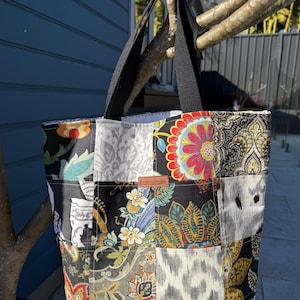 May include: A patchwork tote bag with black handles. The bag features a variety of patterned fabric squares in floral, paisley, and abstract designs. A small brown leather tag is sewn on the front. The bag is hanging from a tree branch.