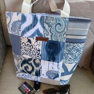 May include: A patchwork tote bag in shades of blue and white, featuring various patterns. It has cream handles and a small brown leather tag. Sunglasses and keys are placed beside the bag.