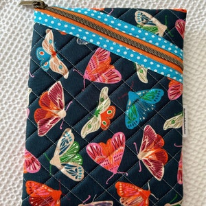 May include: Navy blue quilted pouch with a butterfly design in orange, pink, green, and blue. Features an orange diagonal zip and a blue polka dot trim. White stitching completes the design.