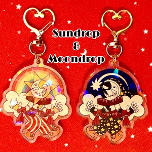 Puede incluir: Dos llaveros iridiscentes con personajes de dibujos animados. El primer llavero presenta un personaje de sol amarillo con un arcoíris y el texto "Sundrop". El segundo llavero presenta un personaje de luna blanca con un cielo estrellado azul y negro y el texto "Moondrop".
