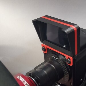 May include: A black camera with a red stripe on the top and a black lens. The camera is mounted on a black stand with a red base.