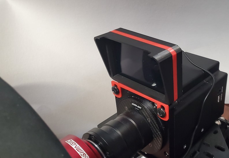 May include: A black camera with a red stripe on the top and a black lens. The camera is mounted on a black stand with a red base.