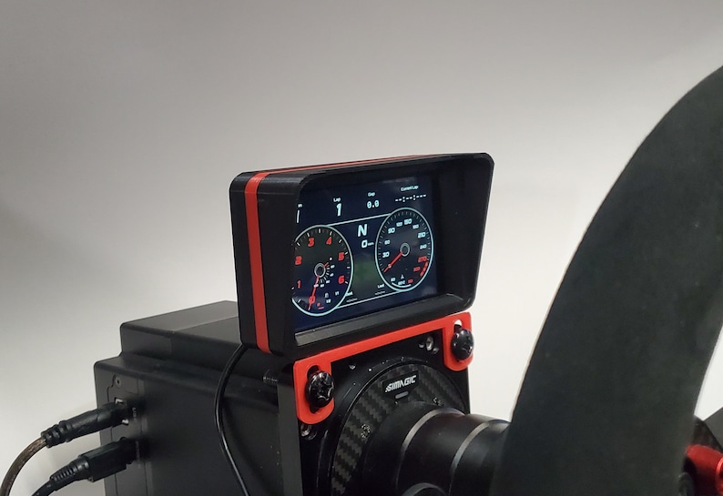 May include: A black racing wheel with a red and black screen mount. The screen displays a digital dashboard with a speedometer, tachometer, and gear indicator. The screen is labeled "SimuLogic".