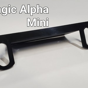 May include: Black plastic bracket with two oval-shaped holes on each side. The bracket is designed to hold a device, such as a phone or tablet, in place. The text "Sim Magic Alpha Mini" is visible in the image.