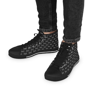 May include: Black high-top sneakers with a repeating gray pattern. The sneakers have black laces, a black toe cap, and a white stripe along the sole. The shoes are worn with black denim jeans.