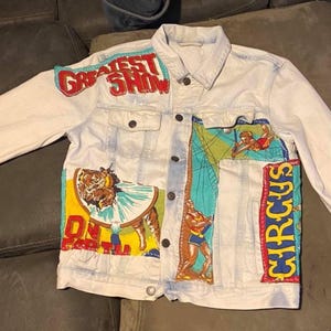 May include: Light wash denim jacket with colourful circus-themed patches. The jacket features the words "GREATEST SHOW" across the chest and "CIRCUS" down the side. Patches depict circus performers and animals.