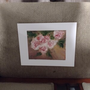 K&ouml;nnte beinhalten: Gerahmtes Gem&auml;lde mit einem Strau&szlig; rosa Rosen und gr&uuml;nen Bl&auml;ttern. Das Kunstwerk ist vor einem hellbraunen Hintergrund und in einem wei&szlig;en Passepartout pr&auml;sentiert. Das Gem&auml;lde wird auf einer gepolsterten Oberfl&auml;che in neutralen T&ouml;nen gezeigt.