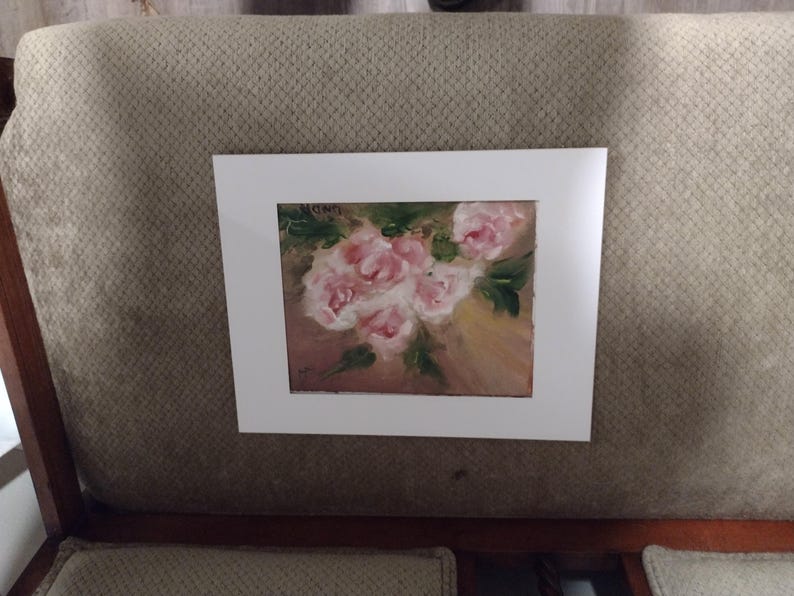 K&ouml;nnte beinhalten: Gerahmtes Gem&auml;lde mit einem Strau&szlig; rosa Rosen und gr&uuml;nen Bl&auml;ttern. Das Kunstwerk ist vor einem hellbraunen Hintergrund und in einem wei&szlig;en Passepartout pr&auml;sentiert. Das Gem&auml;lde wird auf einer gepolsterten Oberfl&auml;che in neutralen T&ouml;nen gezeigt.