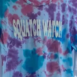 May include: A tie-dye t-shirt with a blue, pink, and purple color scheme. The shirt has the words "Squatch Watch" printed in white on the front.