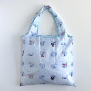 May include: A light blue tote bag featuring a repeating pattern of small cartoon axolotl characters in various poses, including some on skateboards. The fabric is printed with the text "Wooper" and "upah" in a light blue font throughout the design.