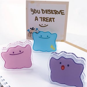 May include: Three acrylic character stands featuring blob-like shapes in pink, blue, and purple. The blue stand holds a square card with the text "YOU DESERVE A TREAT" and a small smiley face. Each character has a simple facial expression.
