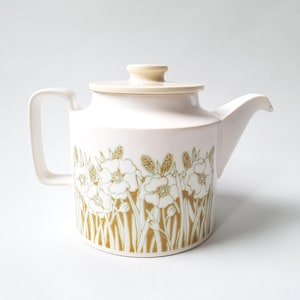May include: A white ceramic teapot with a lid, featuring a floral design in gold and white. The teapot has a curved spout and a handle. The floral pattern includes flowers and tall grass.
