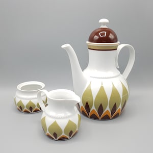 Op de afbeelding: Een witte keramische koffieset met een bruin deksel, bestaande uit een koffiepot, een melkkan en een suikerpot. De set is versierd met een herhalend patroon van olijfgroene, bruine en oranje bladvormen. De koffiepot is ongeveer 25 cm hoog.