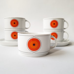 Op de afbeelding: Witte keramische koffiekopjes en schotels met een retro-ontwerp. Elke kop heeft een grote oranje en zwarte cirkelvormige afbeelding. De kopjes zijn gestapeld en gerangschikt op een wit oppervlak, wat een vintage esthetiek oproept.