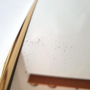 K&ouml;nnte beinhalten: Nahaufnahme eines goldfarbenen, gebogenen Spiegelrahmens vor wei&szlig;em Hintergrund. Die Oberfl&auml;che des Spiegels reflektiert ein verschwommenes Bild, mit kleinen dunklen Flecken auf dem Glas. Der Rahmen hat ein schlankes, modernes Design.