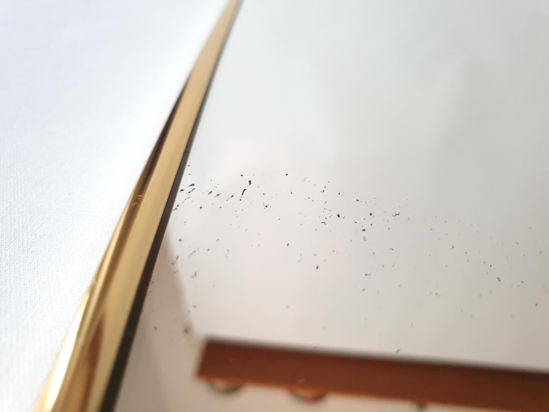 K&ouml;nnte beinhalten: Nahaufnahme eines goldfarbenen, gebogenen Spiegelrahmens vor wei&szlig;em Hintergrund. Die Oberfl&auml;che des Spiegels reflektiert ein verschwommenes Bild, mit kleinen dunklen Flecken auf dem Glas. Der Rahmen hat ein schlankes, modernes Design.