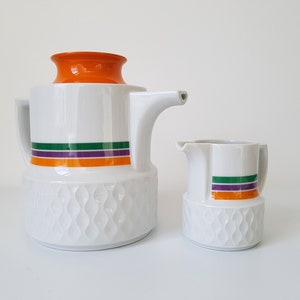Può includere: Set di caffettiera e lattiera in ceramica bianca. La caffettiera ha un coperchio arancione e un design a strisce verdi, viola e arancioni. La lattiera ha un design a strisce abbinato. Entrambi i pezzi hanno un motivo strutturato a forma di onda.