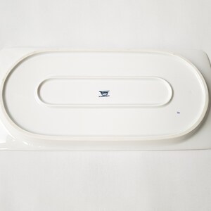 Pu&ograve; includere: Un vassoio da portata rettangolare bianco con angoli arrotondati. Il lato inferiore presenta un'indentazione ovale in rilievo e un logo blu con testo. Il vassoio &egrave; in ceramica o porcellana.