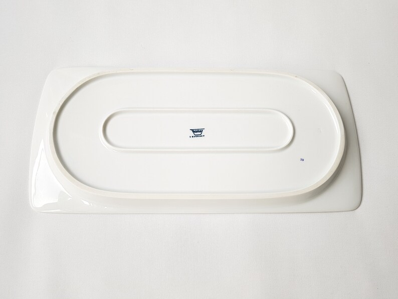 Pu&ograve; includere: Un vassoio da portata rettangolare bianco con angoli arrotondati. Il lato inferiore presenta un'indentazione ovale in rilievo e un logo blu con testo. Il vassoio &egrave; in ceramica o porcellana.