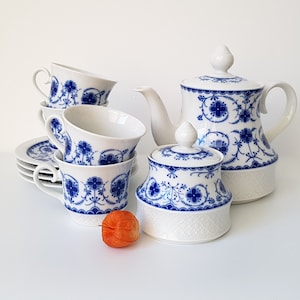 Może przedstawiać: Biały porcelanowy zestaw do herbaty z niebieskimi wzorami kwiatowymi. Zestaw zawiera czajniczek, cukiernicę, filiżanki i spodki. Wzór przedstawia szczegółowe niebieskie kwiaty i liście.