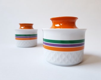 Könnte beinhalten: Zwei weiße Keramikbehälter mit orangefarbenen Deckeln. Jeder Behälter hat horizontale Streifen in Grün, Lila und Orange. Der Boden der Behälter hat ein strukturiertes, sich wiederholendes Muster.