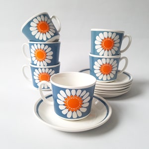 Può includere: Un set di sei tazze in ceramica blu con disegni di margherite bianche e centri arancioni, impilate con piattini abbinati. Le tazze hanno manici bianchi e sono disposte su una superficie bianca. Il set è coordinato.