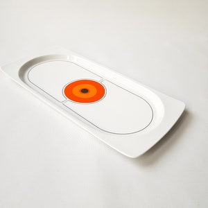 Pu&ograve; includere: Un vassoio da portata rettangolare bianco con un design retr&ograve;. Il vassoio presenta un design ovale centrale con un contorno nero e un motivo circolare di cerchi concentrici arancioni, rossi e marroni. Il vassoio ha un bordo curvo.