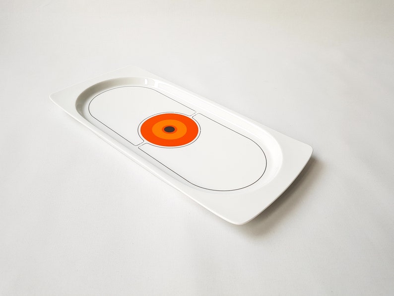 Pu&ograve; includere: Un vassoio da portata rettangolare bianco con un design retr&ograve;. Il vassoio presenta un design ovale centrale con un contorno nero e un motivo circolare di cerchi concentrici arancioni, rossi e marroni. Il vassoio ha un bordo curvo.