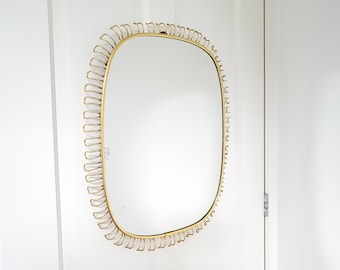 Puede incluir: Un espejo rectangular dorado con esquinas redondeadas. El marco presenta un diseño curvo repetitivo. El espejo está montado en una pared blanca, reflejando la habitación. Una pieza decorativa para la decoración del hogar.