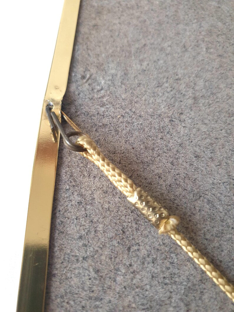 K&ouml;nnte beinhalten: Nahaufnahme eines goldfarbenen Bilderrahmens mit einer geflochtenen goldenen Kordel zum Aufh&auml;ngen. Der Rahmen ist rechteckig und hat ein schlichtes, elegantes Design. Der Hintergrund ist ein ged&auml;mpftes Grau.