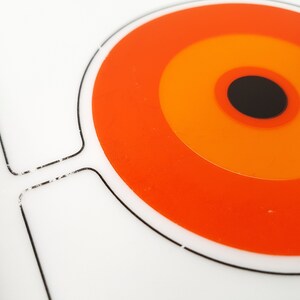 Pu&ograve; includere: Un vassoio da portata bianco in stile retr&ograve; con un audace design circolare arancione e nero. Il vassoio presenta una serie di cerchi concentrici arancioni, giallo-arancio e neri. Il vassoio ha una forma simile a una maniglia sul lato.