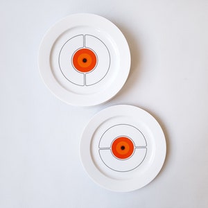 May include: Two white ceramic plates with a retro design. Each plate has a central orange and black bullseye pattern, surrounded by a grey circular outline and divided into four sections. The plates are set against a white background.
