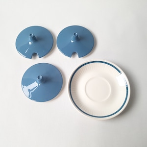 Può includere: Quattro coperchi in ceramica blu con un piccolo poggia cucchiaio e un piattino bianco con bordo blu. Gli oggetti sono disposti su una superficie bianca, suggerendo un ambiente cucina o sala da pranzo.
