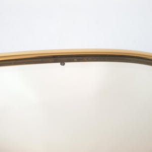 K&ouml;nnte beinhalten: Nahaufnahme eines goldfarbenen, oval geformten Spiegelrahmens. Der Rahmen hat ein d&uuml;nnes, metallisches Aussehen mit einem leicht gebogenen Design. Der Hintergrund ist eine schlichte wei&szlig;e Oberfl&auml;che.