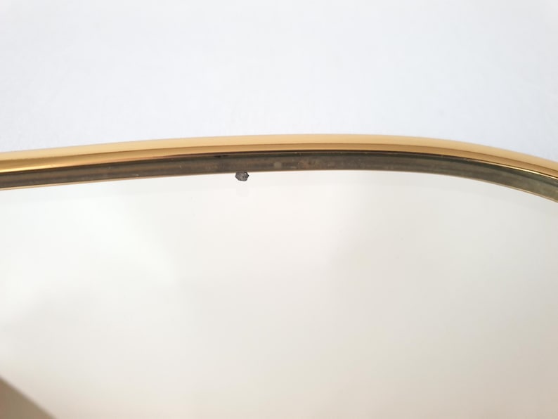 K&ouml;nnte beinhalten: Nahaufnahme eines goldfarbenen, oval geformten Spiegelrahmens. Der Rahmen hat ein d&uuml;nnes, metallisches Aussehen mit einem leicht gebogenen Design. Der Hintergrund ist eine schlichte wei&szlig;e Oberfl&auml;che.