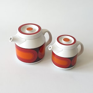 Puede incluir: Juego de té retro de dos piezas. La tetera y la jarra de crema son blancas con un diseño geométrico rojo, naranja y marrón. Cada pieza tiene una tapa blanca con un acento naranja circular. La tetera tiene un asa y un pico.