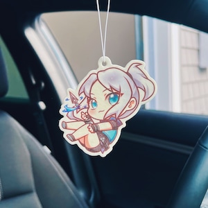 May include: A white car air freshener featuring a cartoon illustration of a young woman with blue eyes and white hair. She is wearing a blue and white outfit and holding a blue weapon.