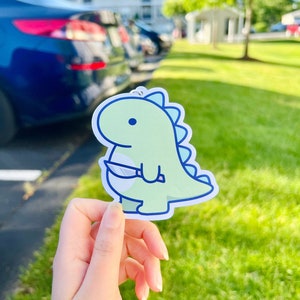 May include: A green dinosaur cartoon character holding a knife. The dinosaur is light green with a blue outline and has a light blue eye. The knife is white with a black handle.