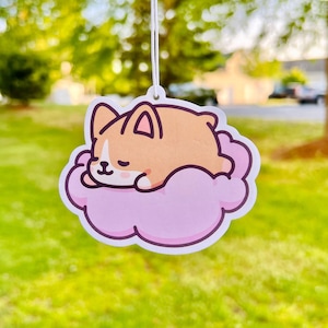 May include: A car air freshener featuring a cartoon corgi dog sleeping on a pink cloud.