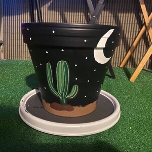 May include: Black terracotta planter with a white crescent moon and a green cactus painted on it. The planter is sitting on a white saucer.