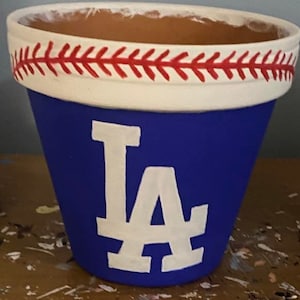 May include: A blue terracotta planter with a white baseball stitch design around the top rim. The planter is painted with a white "LA" logo.