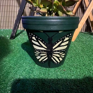 May include: A dark green terracotta planter with a painted white butterfly design. The butterfly has black markings and is centered on the front of the planter.