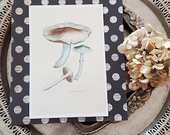 May include: A watercolor illustration of several mushrooms in shades of red, green, and blue. The artwork is on a white card, layered over a black patterned paper with white polka dots and text. The arrangement is on a silver tray.