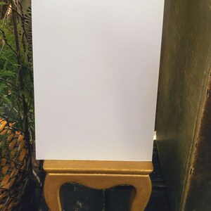 May include: A blank white card stands on a small, ornate gold-colored wooden stand. The stand has curved legs and a rectangular top. A vintage book with a green cover is to the right of the card. The background includes a decorative silver tray and greenery.