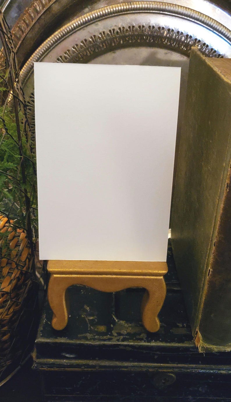 May include: A blank white card stands on a small, ornate gold-colored wooden stand. The stand has curved legs and a rectangular top. A vintage book with a green cover is to the right of the card. The background includes a decorative silver tray and greenery.