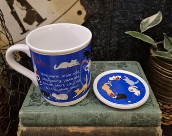 May include: A white ceramic mug and matching circular lid featuring a blue background with illustrations of various cats playing with red yarn. The mug displays the text: Cats... blink warm, stare cold, play young, yawn old, crouch short, strut tall, act big, sleep small.