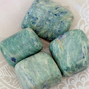 May include: Four green and blue tumbled stones. The stones have a smooth, rounded shape and a marbled appearance. The colours vary in intensity, creating a natural, earthy aesthetic. The stones are arranged on a white surface.
