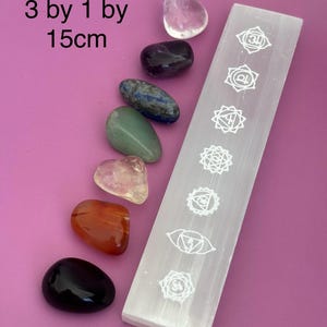 May include: A selection of polished gemstones in shades of pink, purple, green, and orange, displayed with a white selenite wand. The wand is etched with chakra symbols. The image text reads "3 by 1 by 15cm".